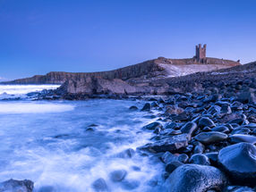 Dunstanburgh Castles stands above shiny boulders at Embleton Bay during the Blue Hour, on the Northumberland Coast