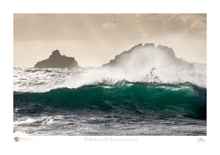 A turquoise wave, backlit by the sun breaks off the coast of Cape Cornwall, with the two small islands of the Brisons lying offshore and partially obscured by spray