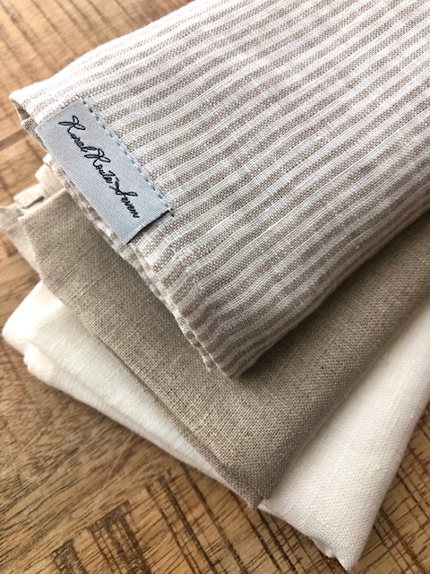 close up of three folded tea towels in White with Natural Flax Stripes, Natural Flax, and Off-White Stonewashed Linen Tea Towel with Rural Route Seven logo brand label, resting on distressed antique pine tabletop