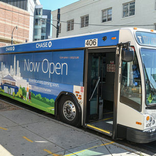 Blue-and-white city bus with "Now Open" ad, parked on urban street. Background shows modern and brick buildings. Bus number 406.