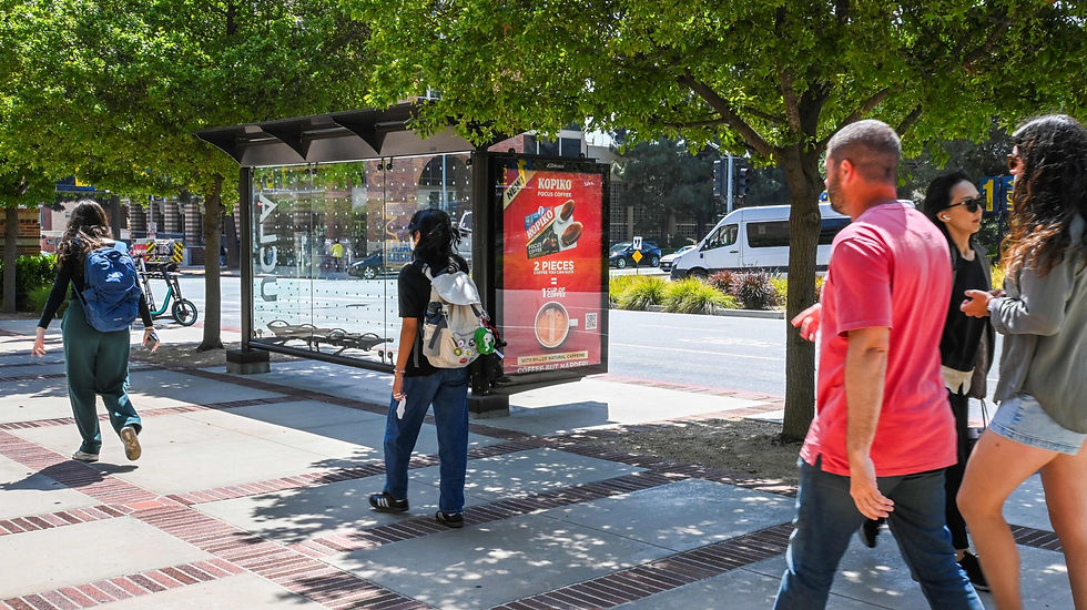 People walk by a bus stop under trees. A prominent red KOPIKO coffee ad is visible. Bright, sunny day with a relaxed atmosphere.
