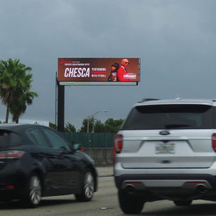 Cars drive past a billboard on a highway promoting Chesca performing with Pitbull. Overcast sky and palm trees in the background.