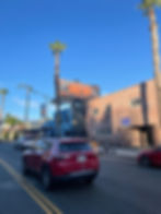 Red Jeep on urban street with palm trees, brick building, "BLOCK PARTY" billboard, and "For Lease" sign. Clear blue sky.
