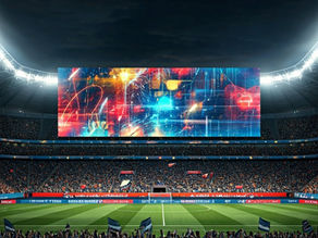 Crowd in a packed night stadium waves flags beneath a giant colorful screen, with bright floodlights over the soccer field.