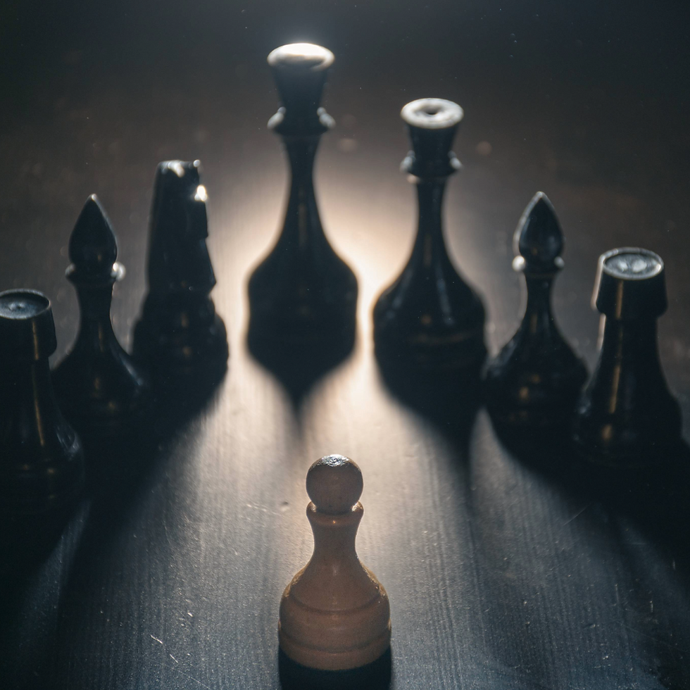 A white pawn stands alone facing a row of black chess pieces on a dark surface, illuminated dramatically from behind, creating a tense mood.