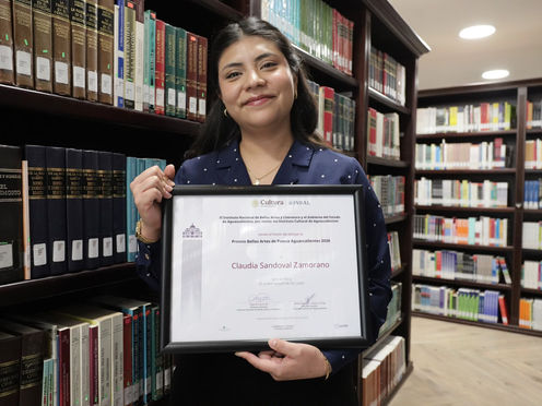 La poeta Claudia Sandoval Zamorano sosteniendo su galardón literario frente a un estante lleno de libros.
