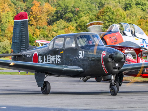 A classic military-style aircraft with Japanese markings taxis on an airfield. Another aircraft is visible behind, surrounded by vibrant autumn trees.