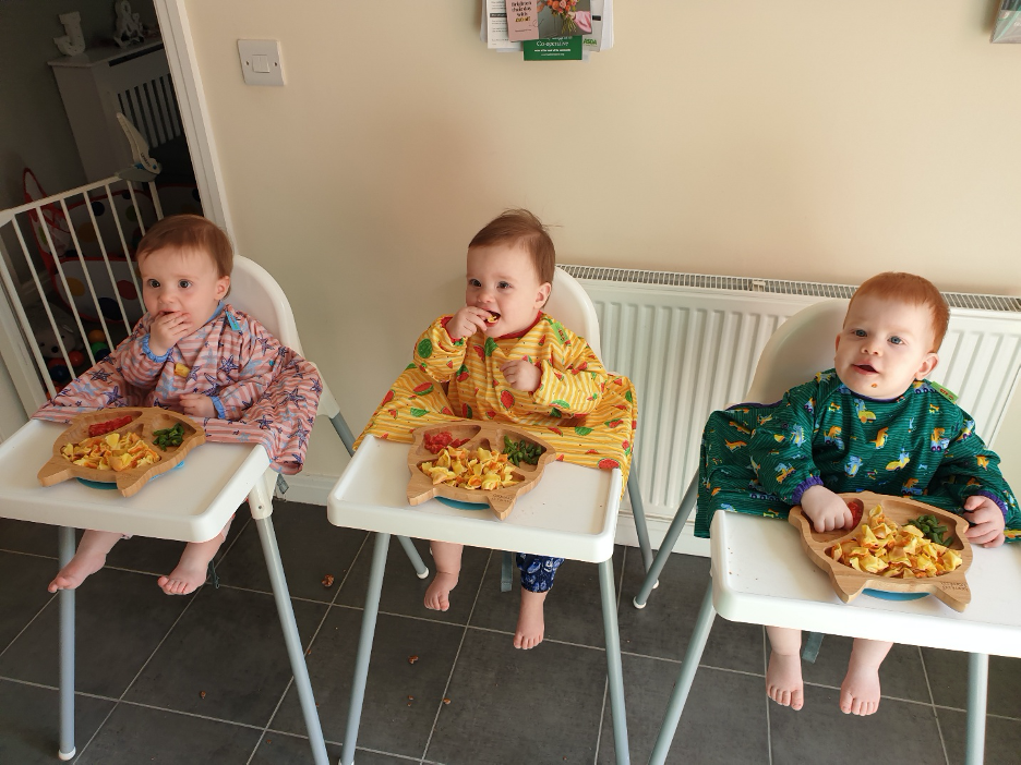 triplets sat in high chairs wearing bibs eating finger foods