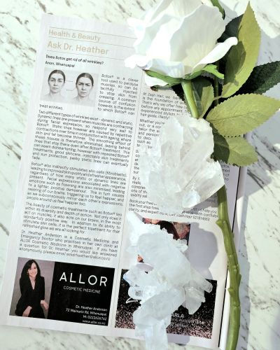Magazine open to an article titled "Ask Dr. Heather" about Botox, with diagrams. A white rose and crystals rest on the page, set on marble.