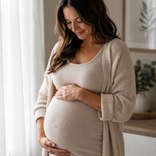 Pregnant woman in beige dress and cardigan gently holding her belly in a soft-lit home, smiling peacefully.