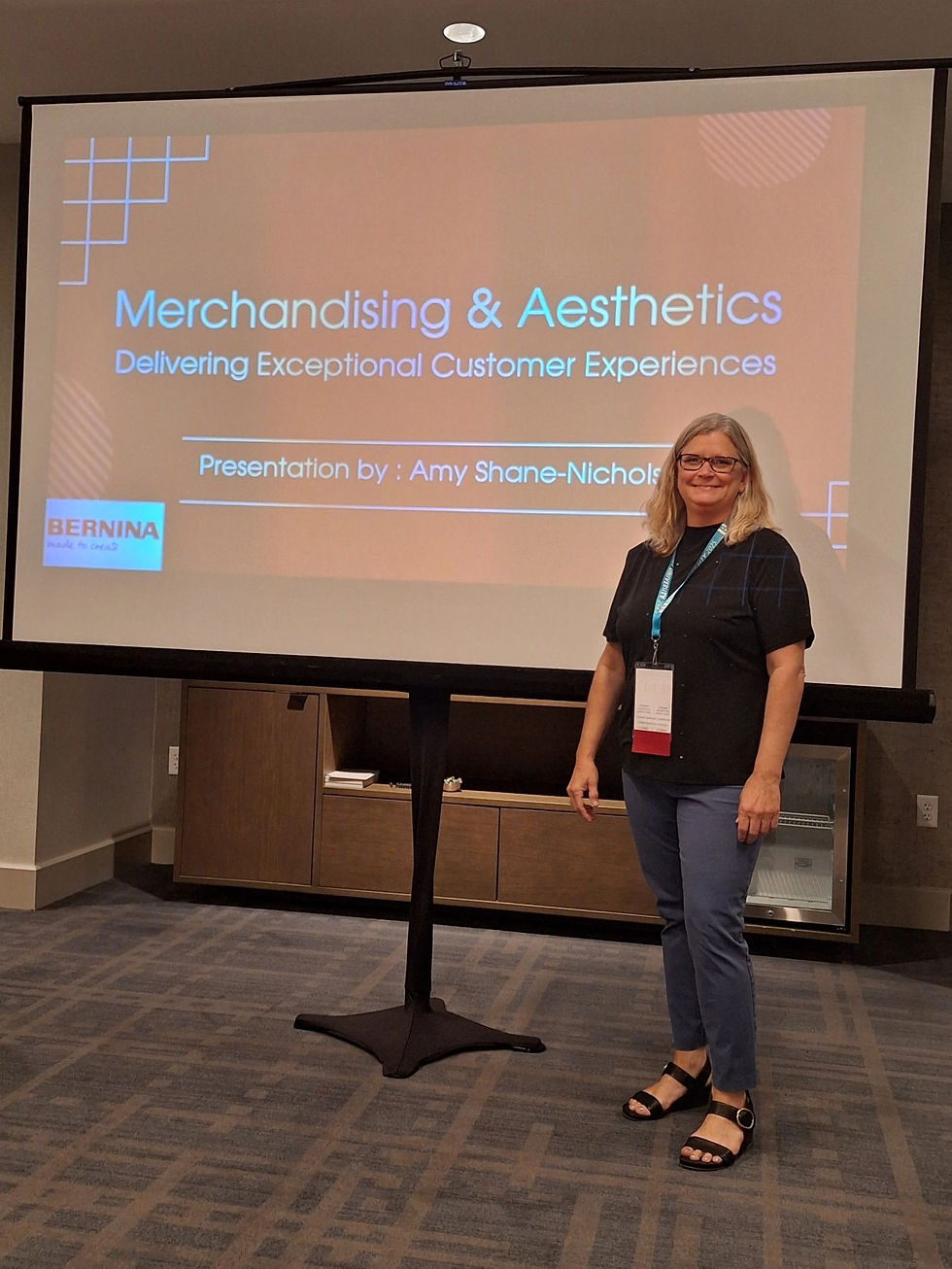 Woman smiling beside presentation screen titled Merchandising & Aesthetics. Text and grid pattern visible. Professional setting.