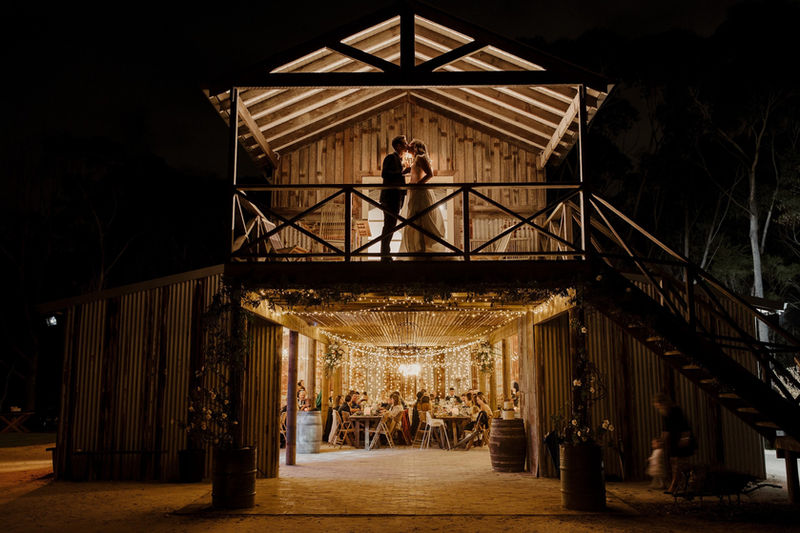Couple on the Loft balcony at night with their guests celebrating in the fairy lit Barn below, The Stables of Somersby