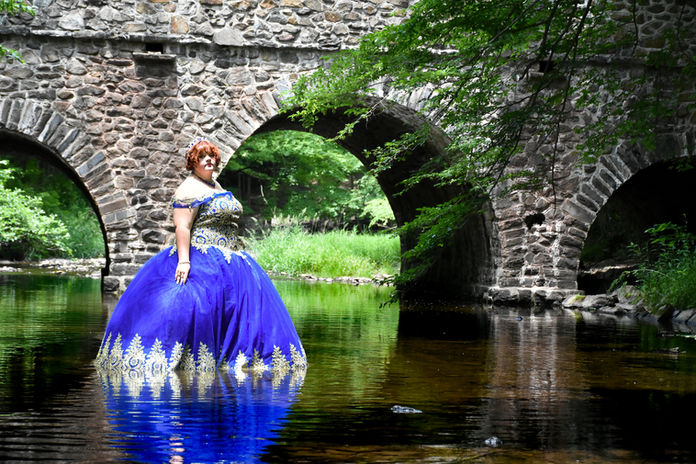 Senior portrait featuring beautiful girl in quinceanera dress by an arched bridge with reflection in stream.