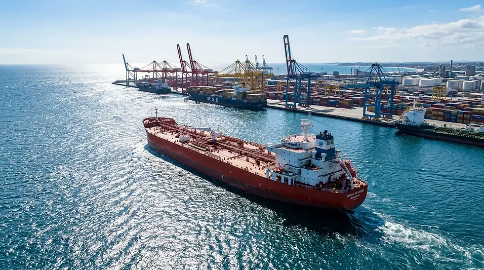 Oil tanker crossing a sunlit ocean near a busy port with cranes.