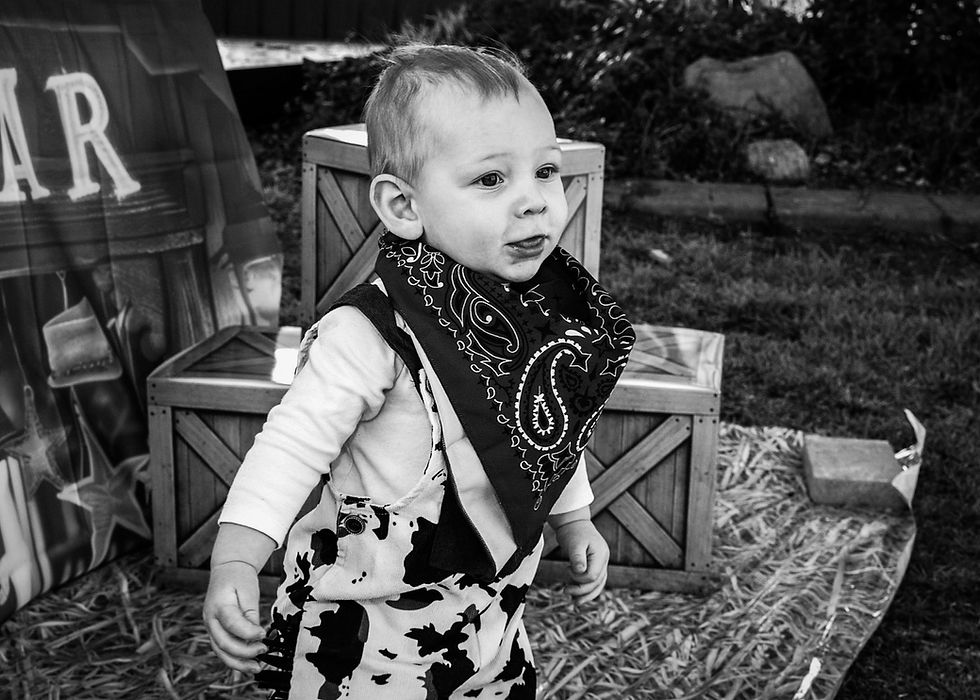 Black-and-white photo of a toddler in a cow-print outfit and bandana standing on hay beside wooden crates outdoors.
