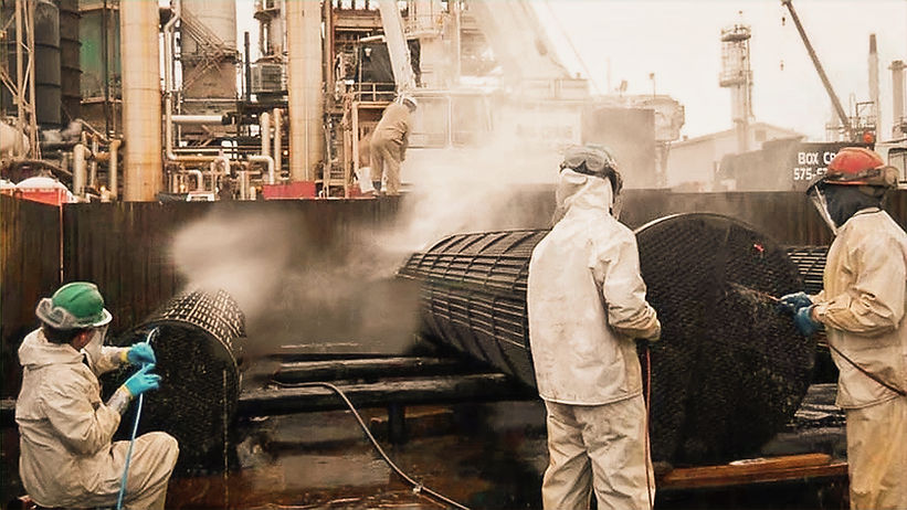 Workers in white suits manage steam and spray from high-pressure water cleaning of large heat exchangers at an industrial site