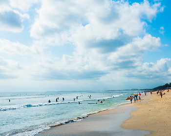 People swim and surf in the ocean under a partly cloudy sky. A sandy beach on the right is dotted with beachgoers, creating a lively scene.