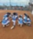 Youth softball team in blue-and-white jerseys kneels in a pregame prayer on a dirt field, with spectators and fences behind.