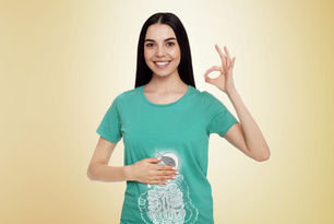 Smiling woman in green shirt with digestive system graphic, making an "OK" gesture against a beige background, exuding positivity.