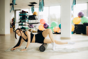 Woman in gym attire using a foam roller on her outer thigh on a mat in a bright studio, surrounded by colorful exercise balls and equipment. Calm mood.