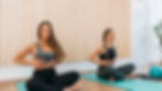 Two women practicing diaphragmic breathing on yoga mats, eyes closed, in a serene room with wood paneling.