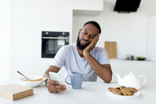 Man in white shirt, tired, doomscrolling at table with cereal, blue mug, and cookies. Bright kitchen background, lethargic mood.