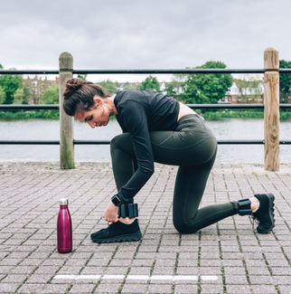 Woman in workout gear kneels to tie shoe by a river. Wearing ankle weights. Pink water bottle nearby. Cloudy sky, pensive mood.
