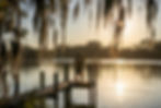 Happy couple standing arm-in-arm on a private wooden dock at golden hour overlooking the calm St. Johns River in Putnam County Florida, with Spanish moss hanging from trees