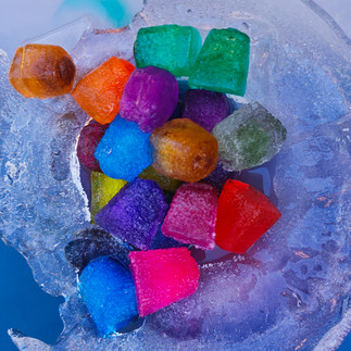 Ein Kind hält mit einer schwarzen Zange einen roten, gefrorenen Eiswürfel über eine große, runde Eisschale, in der bunte Eiswürfel in verschiedenen Farben liegen. Im Hintergrund sind weitere bunte Eiswürfel und Kinderhände zu sehen