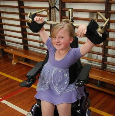 A girl in a ballet outfit, using a powered arm support attached to her wheelchair to lift arms above her head..