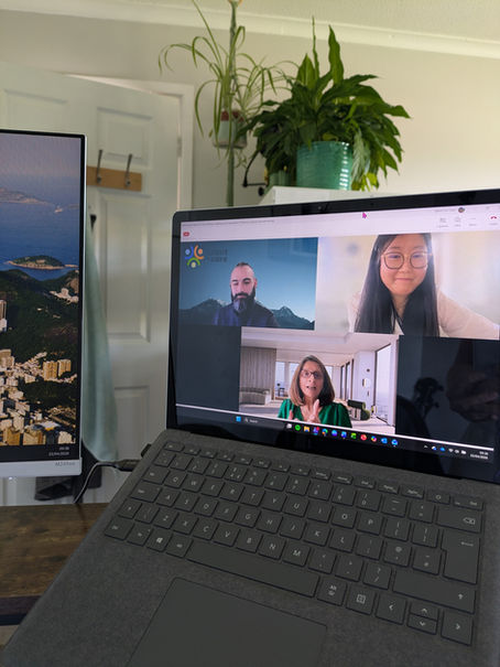 A laptop screen showing an active Webinar call, with three participants visible: a man with a beard against a mountain backdrop (Arran Daly), a woman with glasses and long dark hair in a white top (Dr Megan Peng), and a woman with shoulder-length hair in a green top, gesturing mid-conversation (Dr Daljit Hothi). The Talent for Care logo is visible in the top-left of the screen. A second monitor showing a coastal landscape is visible to the left. The photo is taken from behind the laptop in a home workspace setting, with a large plant in the background.
