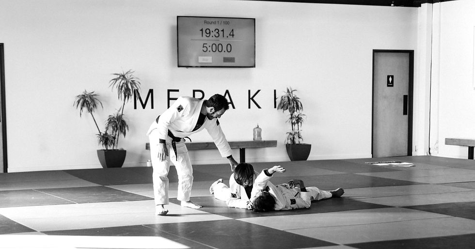 Martial arts coach assists two students on mats in a bright studio. "MERAKI" on wall. Timer displays "19:31.4" and "5:00.0".