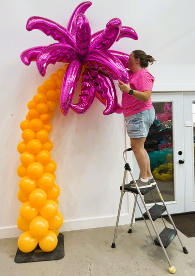 Balloon artist installing a pink foil palm tree topper on an orange balloon column.