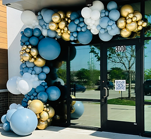 Outdoor balloon garland installation framing a storefront entrance with a mix of matte blue, gold, black, and white balloons in varying sizes, designed by North Texas Balloons