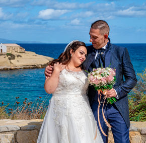 Sposi abbracciati con bouquet davanti al mare blu.