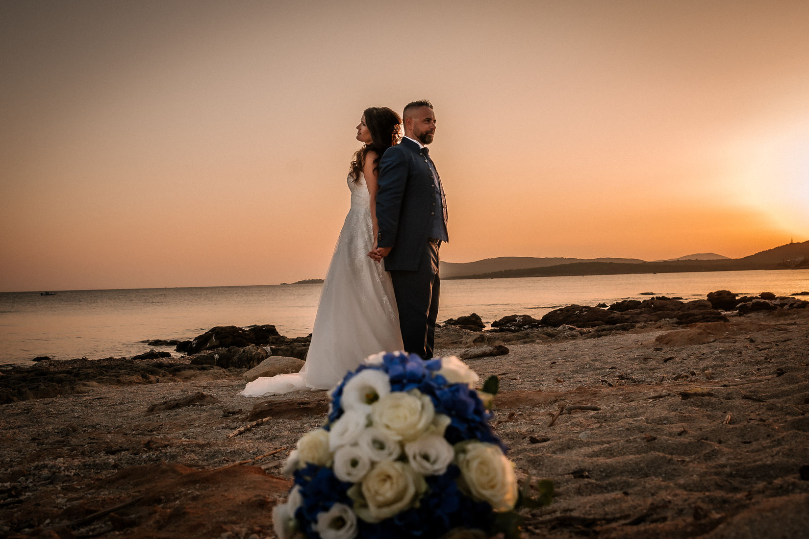 Sposi al tramonto su spiaggia, bouquet in primo piano.