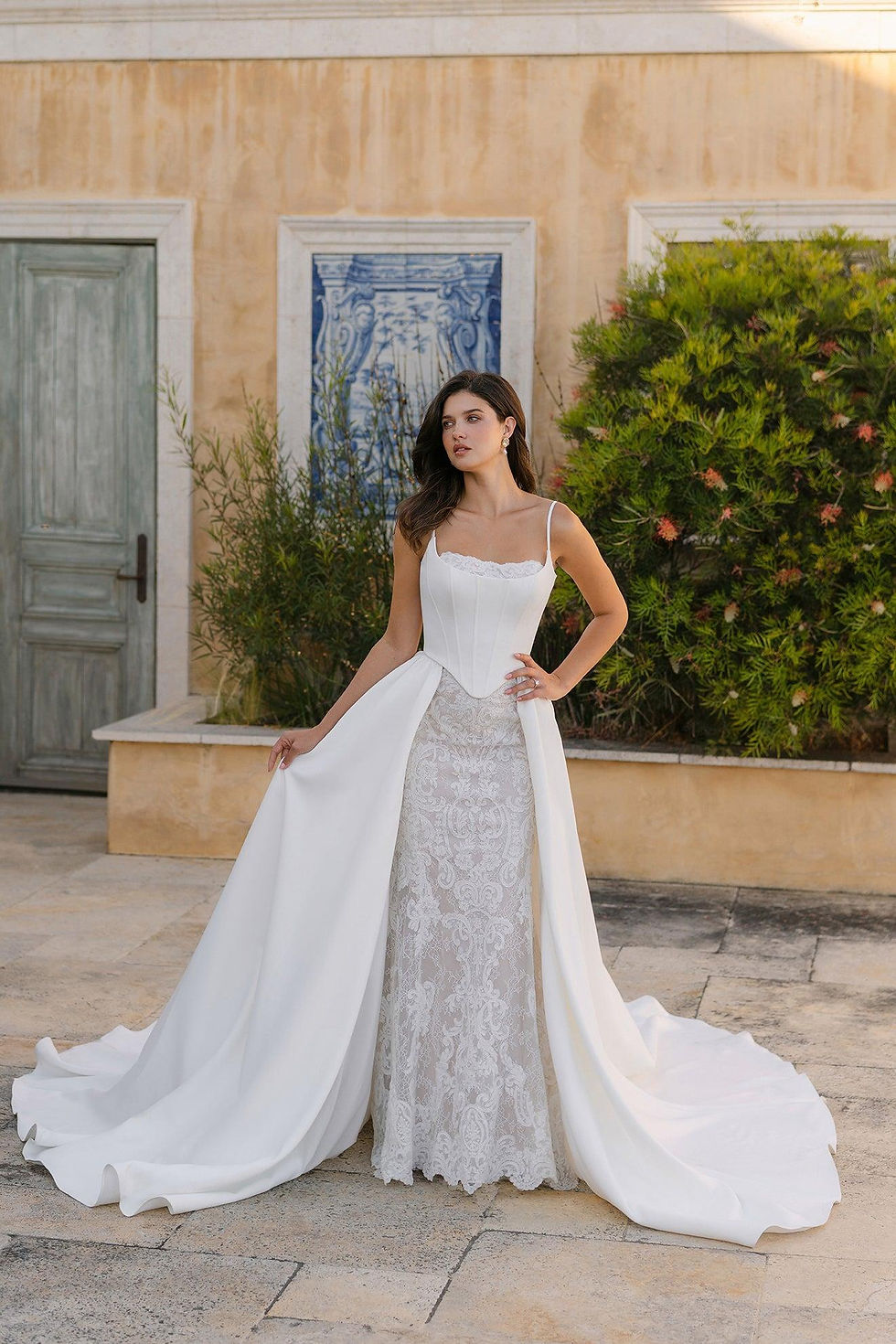 Woman in a white lace bridal gown with long train poses outdoors beside a stucco wall, green shrubs, and a blue panel.