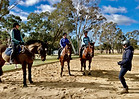 Three mature female riders sit on bay horses in a semi circle. The horses are on a sand arena and in front of the semi circle is a female coach in navy blue demonstrating a riding position.