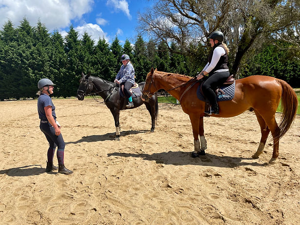 Two woman sit on horses speaking to a riding coach. All three are laughing.