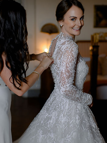 bride smiling as a bridesmaid fastens the delicate lace details of her wedding gown, illuminated by soft natural light