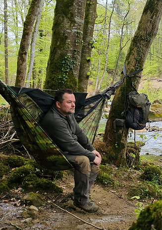 Man rests in a hammock beside a forest stream, backpack hanging from a tree during a quiet wilderness break.