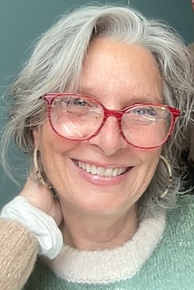 An image of Catriona O'Curry: a woman with pinkish white skin, crystal-blue eyes, white and grey hair in a swoop over her forehead, large red glasses, hoop earrings, and a welcoming smile showing her front teeth. Her hand holds her neck, she wears a pretty knit sweater, and she stands before a light blue wall.