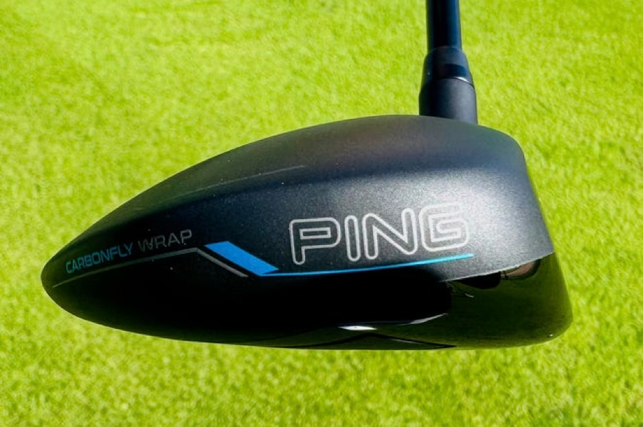 Close-up of a black PING golf driver head with Carbonfly Wrap on bright green grass, showing blue accents and a sleek look