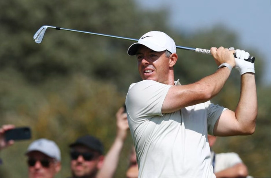 Golfer in a white cap and shirt follows through on a swing at a sunny course, with blurred spectators in the background.