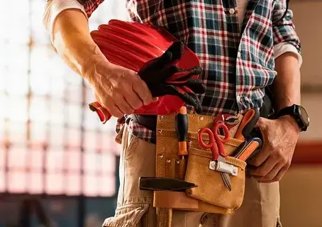Close-up of a handyman wearing a tool belt and holding a red hard hat.