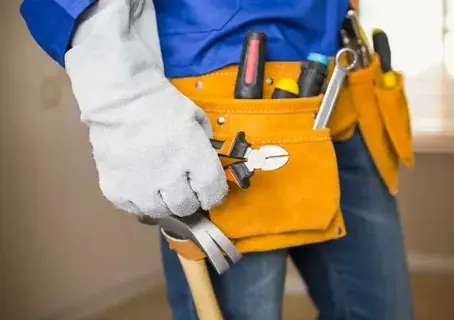A handyman wearing a tool belt filled with various tools, holding pliers and a hammer.