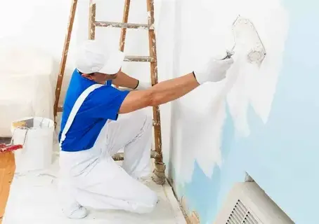 A professional painter wearing white overalls and a blue shirt repairing and painting a wall in a home interior.