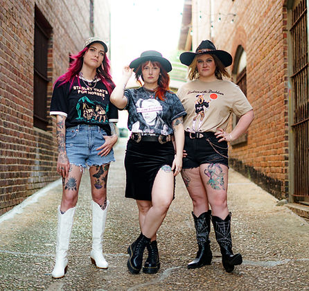 Three women in a narrow brick alley. They wear stylish hats, graphic tees, and boots. The mood is bold and trendy.