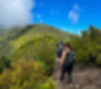 Two hikers on a narrow trail towards Pico Ruivo, having left the forest behind. The path offers breathtaking views of green mountains and a bright blue sky with scattered clouds. The hikers have a few scratches but are enjoying the beautiful scenery.