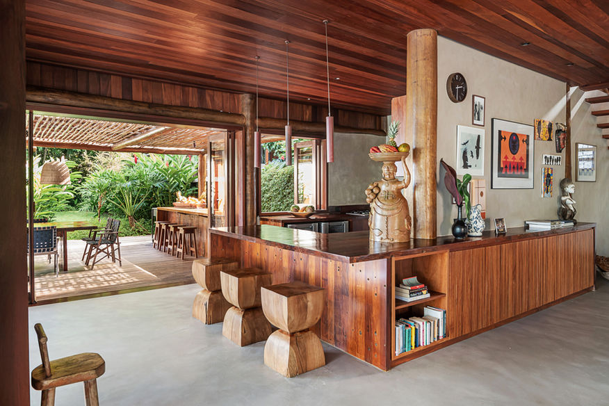 Cozinha com bancada de madeira, banquetas e prateleiras, integrada ao ambiente de uma Barracuda Villa em Itacaré, Bahia.